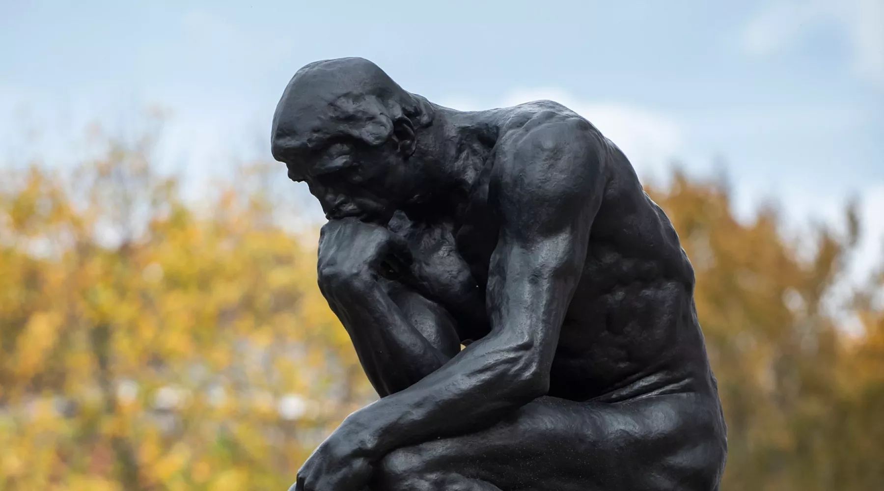 Side profile of The Thinker statue by Auguste Rodin outside of Grawemeyer Hall with fall foliage in the background