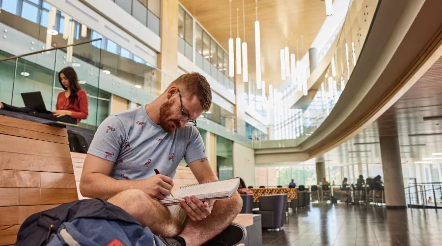 Student sitting with their legs crossed on the wooden platform benches in the Belknap Academic Building reading an academic textbook.