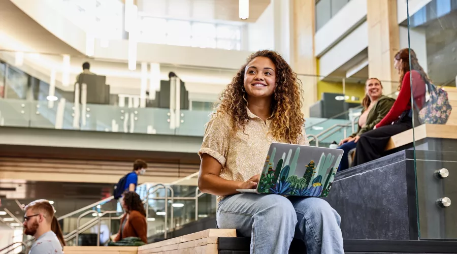 Student recognizes another student walk into the Belknap Academic Building as she is typing on her laptop and sitting on the wooden platform benches at the entrance of the BAB.