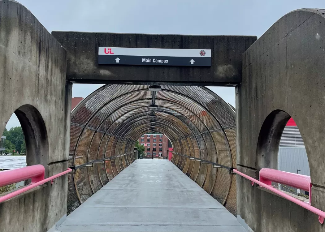 Perspective focusing down a walkway from UofL's parking garage to campus. Signage above circular walkway reads "Main Campus" with arrows pointing ahead. Campus can be seen down the walkway.