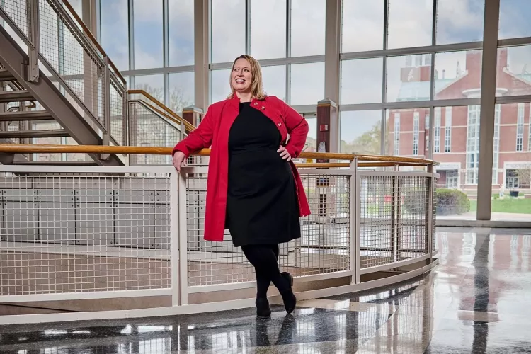A smiling woman in a red jacket and black dress poses against a stairway railing.